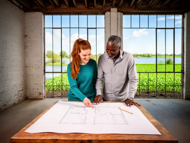 Two business partners smiling over a floor-plan drawing in an empty brick warehouse with lake and crop views